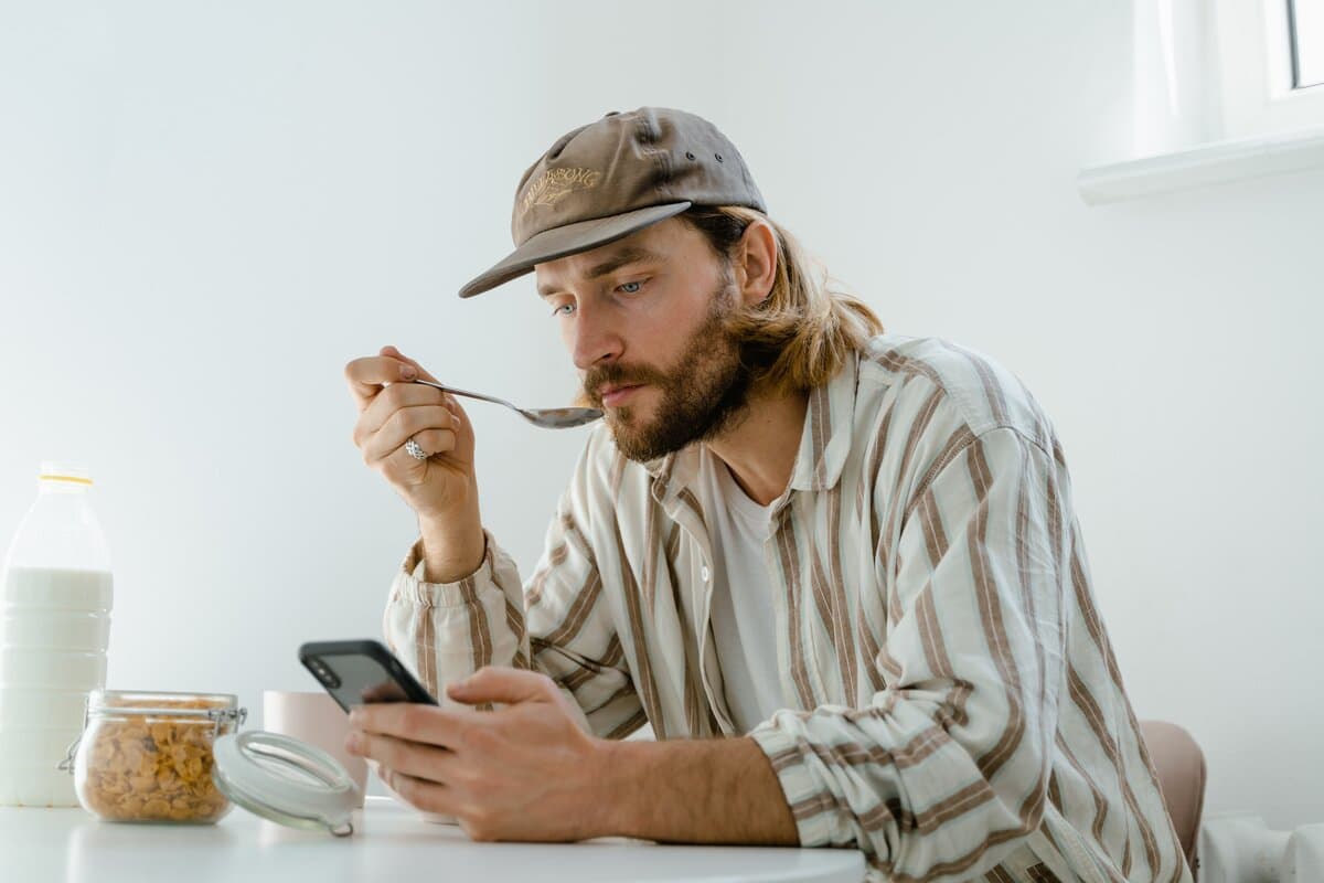 A man in a casual striped shirt and cap looking at e-learning on his phone while eating lunch