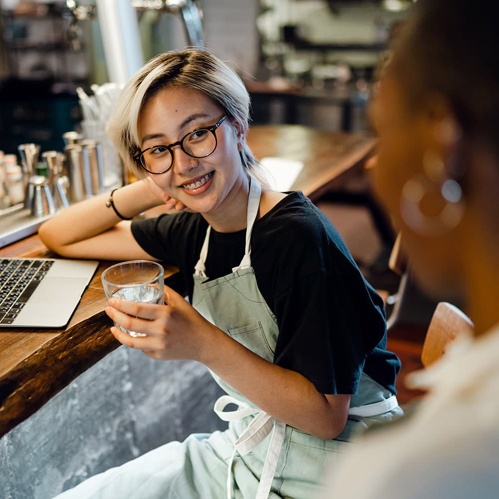 Two women in a restaurant setting having a conversation. The one in focus is sitting at a bar counter with an open laptop, holding a glass of water.