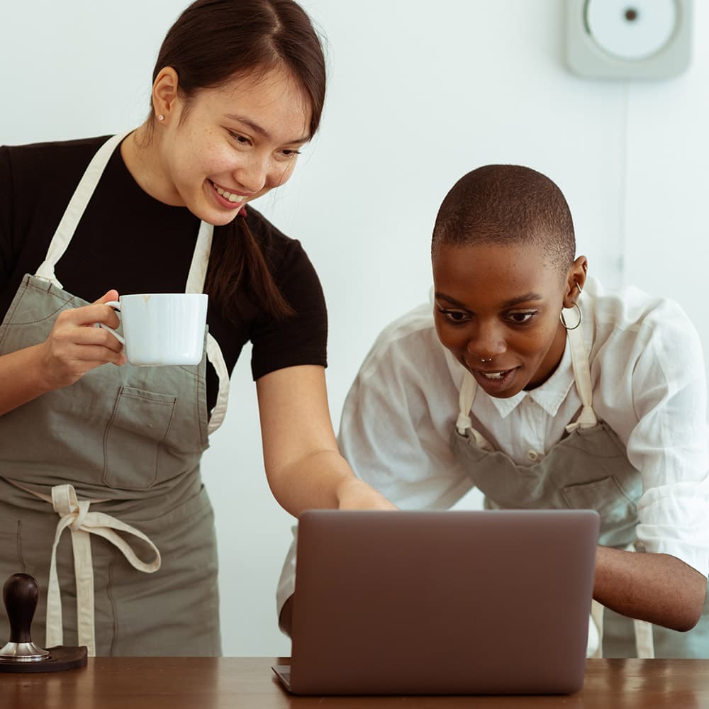 A cafe owner with a coffee cup in hand showing a staff member something on a laptop.
